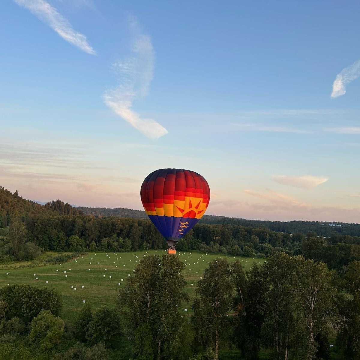 A hot air balloon with the balloon decorated to look like a sunset floating above a lush green landscape. the sky is bright and mostly clear with little white clouds
